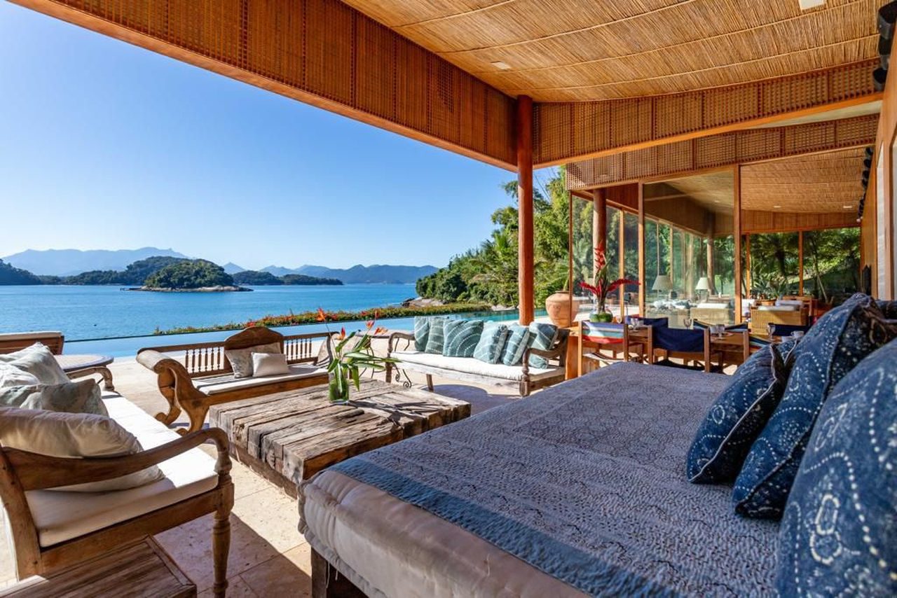 Sala de estar social coberta, com daybed e sofás, totalmente aberta para o mar da enseada e as ilhas ao fundo