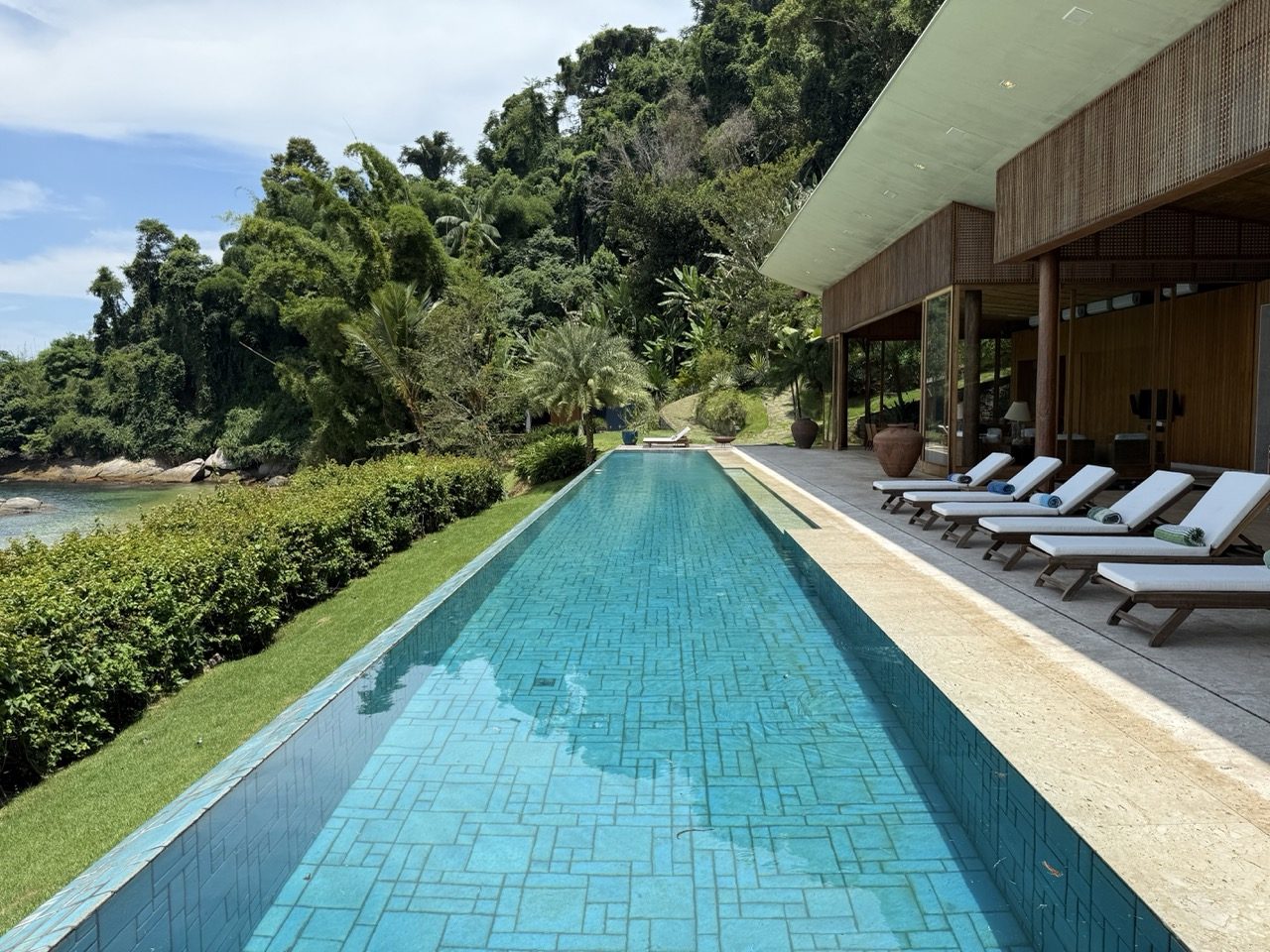 A piscina de borda infinita de 36 metros corre ao longo do deck, abraçada pela mata nativa, com a praia e o mar da enseada ao fundo